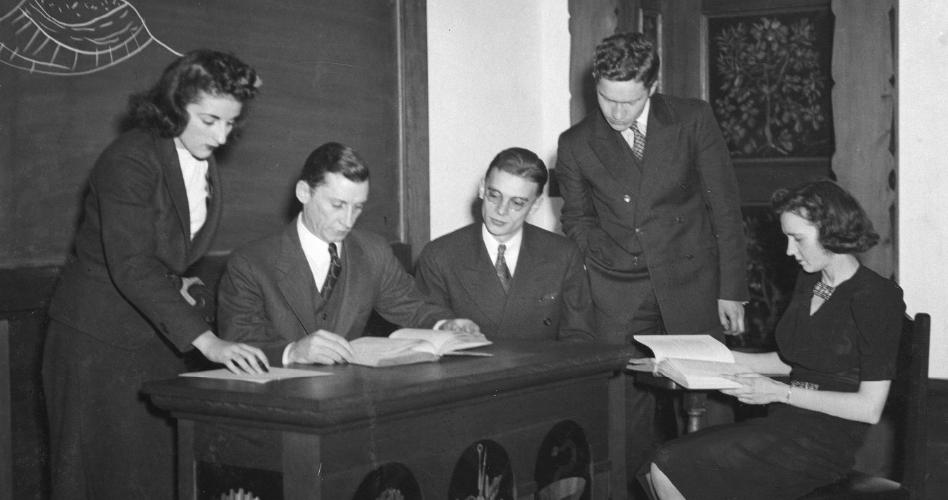 Dr. Richard E. Sherill sits at a desk with a book, while four students look on.