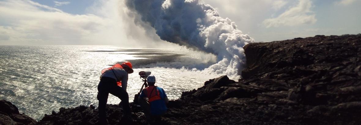Two people stand on jagged rock studying a smoke plume.