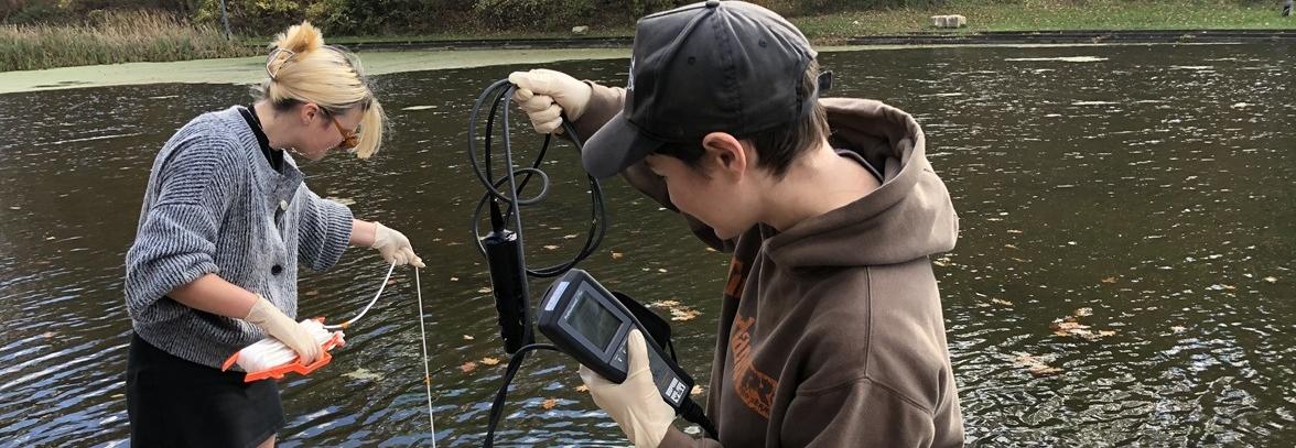 Two students stand next to a body of water with testing equipment.